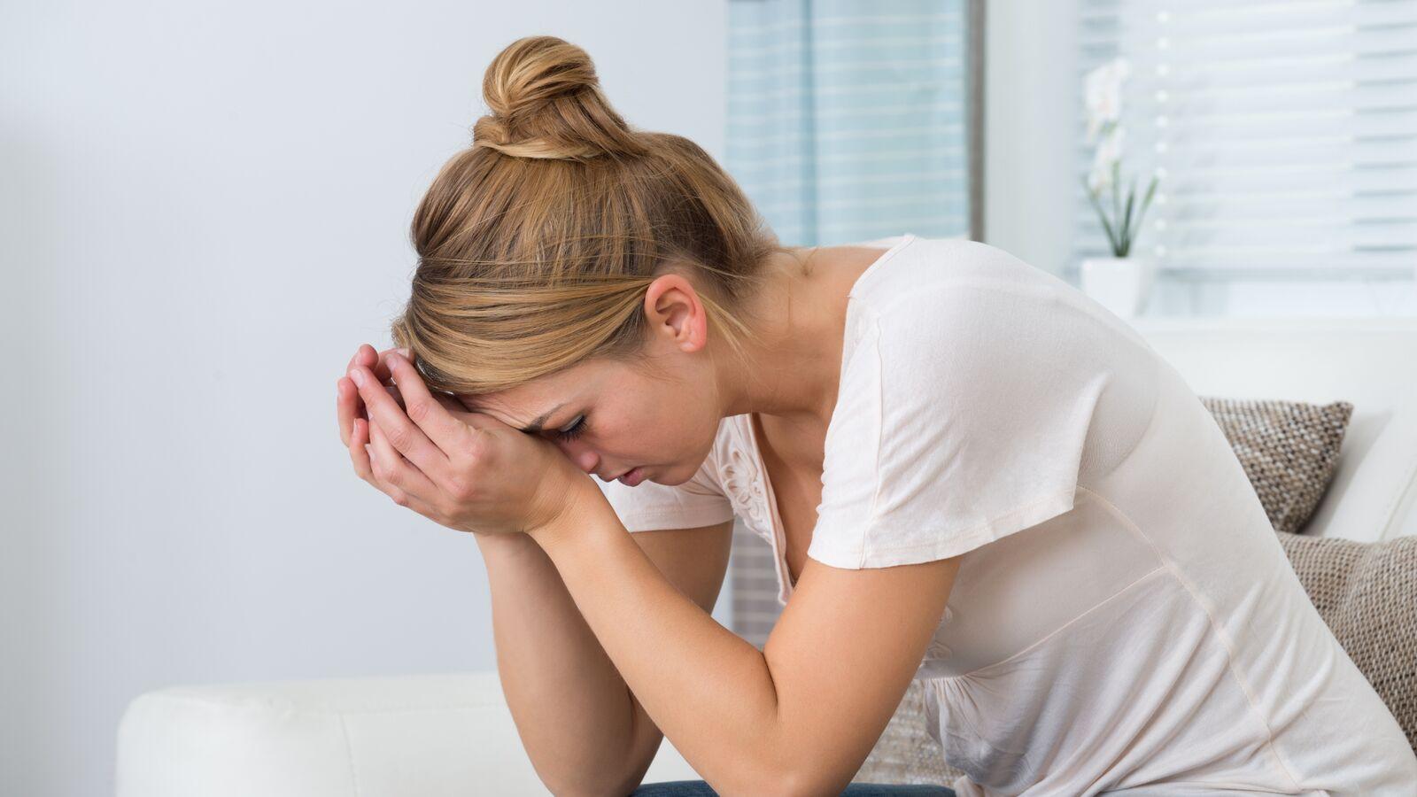 Jeune femme assise sur un canapé, tête baissée et front appuyé sur ses mains jointes, en proie au stress